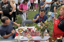 Tłum ludzi i dwoje mężczyzn kosztujących danie przy stole. Obok nich wnikliwie przyglądająca się kobieta 